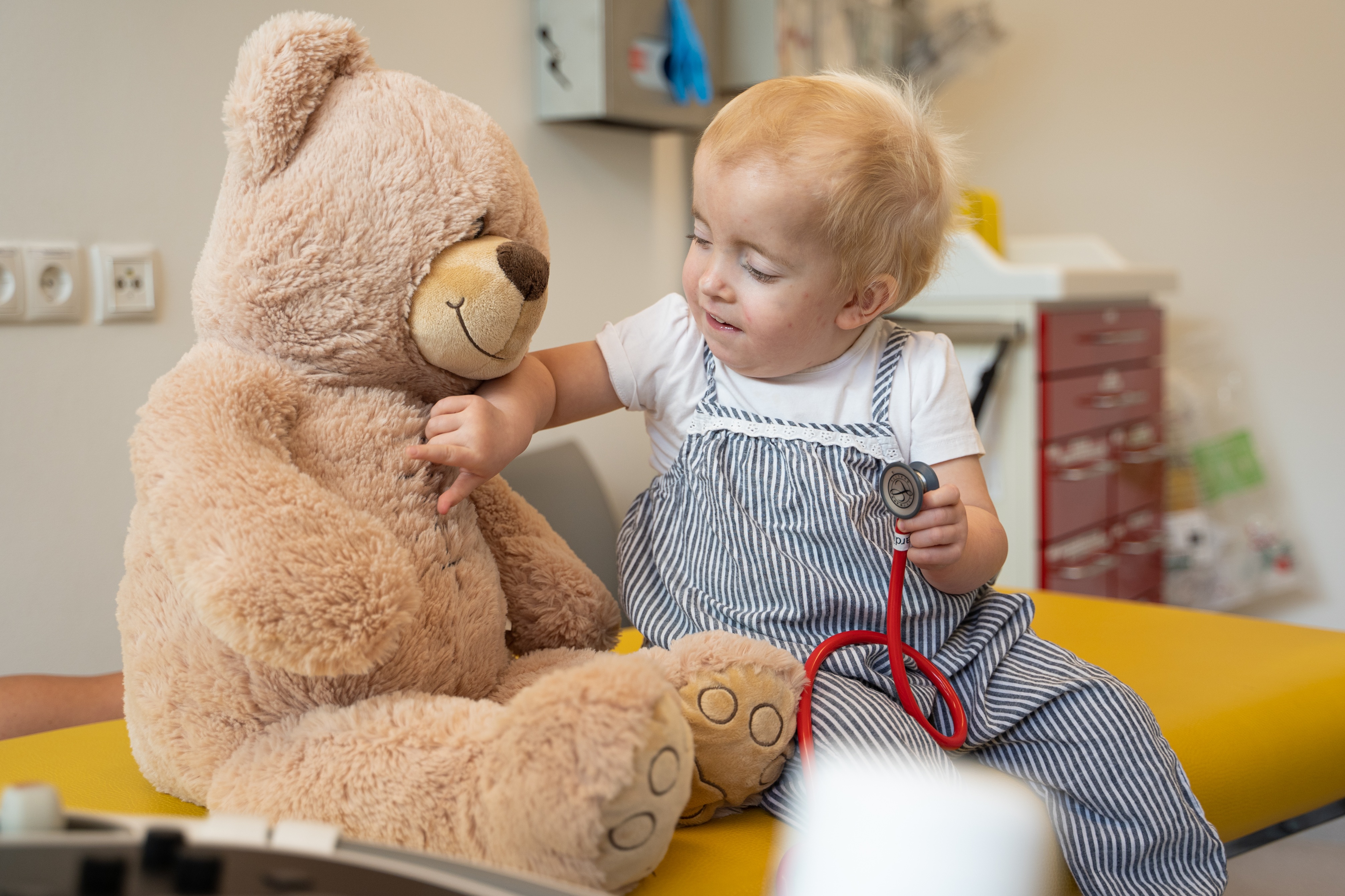 Kind spielt auf Untersuchungsliege mit großem Teddy