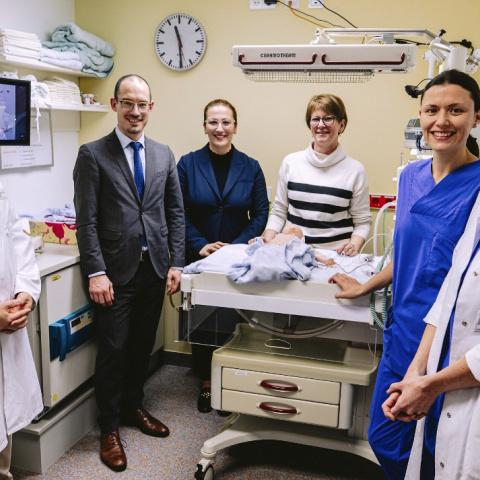 Die Gruppe steht um die Behandlungsliege mit Babypuppe. Im Hintergrund sieht man den zugeschalteten Arzt am Bildschirm.