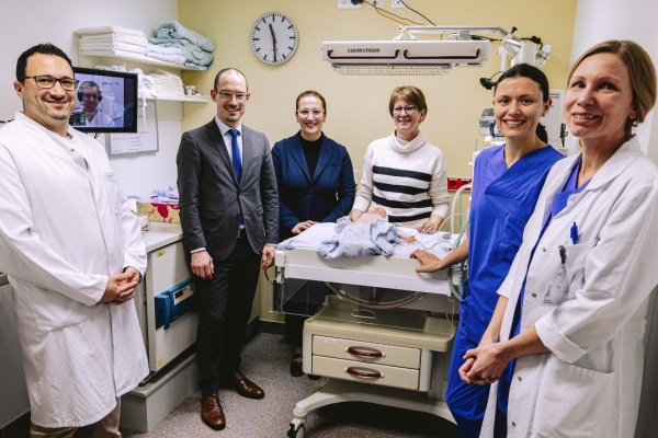 Die Gruppe steht um die Behandlungsliege mit Babypuppe. Im Hintergrund sieht man den Bildschirm mit dem Oberarzt aus dem Olgahospital.