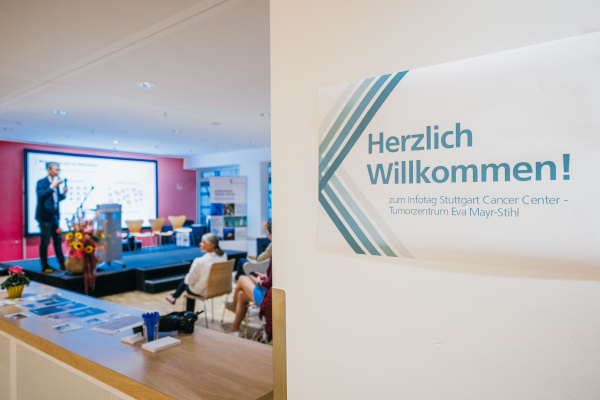 Schild mit "Herzliche Willkommen zum Infotag Stuttgart Cancer Center" und Bühne mit Sprecher im Hintergrund