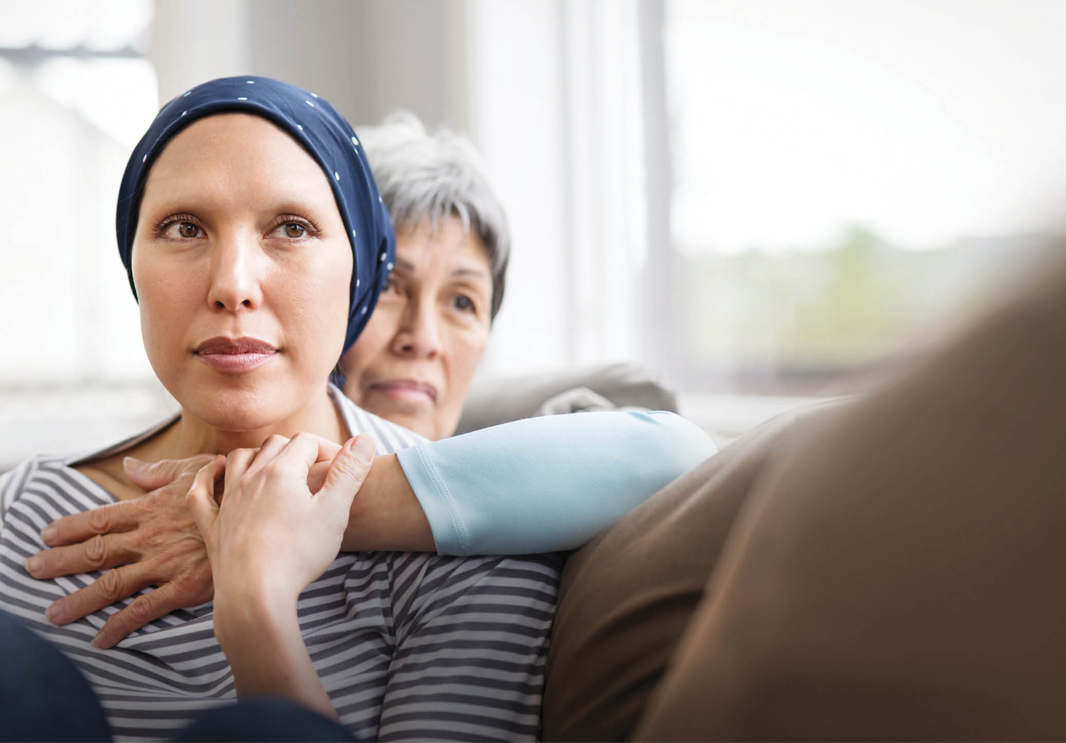 Asian woman in 60s embracing her mid-30s daughter who is fighting cancer and is wearing a headcovering  They are seated on a couch and have contemplative expressions 